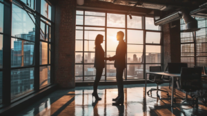 Manager and new employee shake hands in a modern fintech office. The sun is low in the sky and a city is visible through the window.
